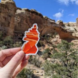 May include: A red and orange sticker of a rock formation in a desert landscape. The sticker is held in a hand in front of a natural rock arch.