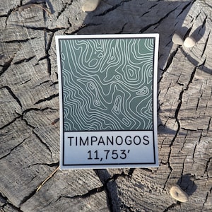 May include: A white sticker with a green topographic map design and the text "TIMPANOGOS 11,753'" on a wooden surface.