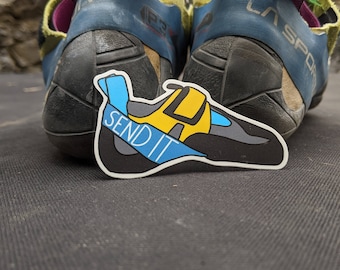 May include: A pair of blue and green climbing shoes with purple straps. The shoes are on a black surface with a sticker that says "Send It" on it.