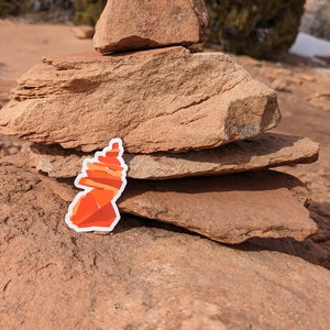 May include: A red and orange rock formation sticker placed on a stack of brown rocks in a desert landscape.