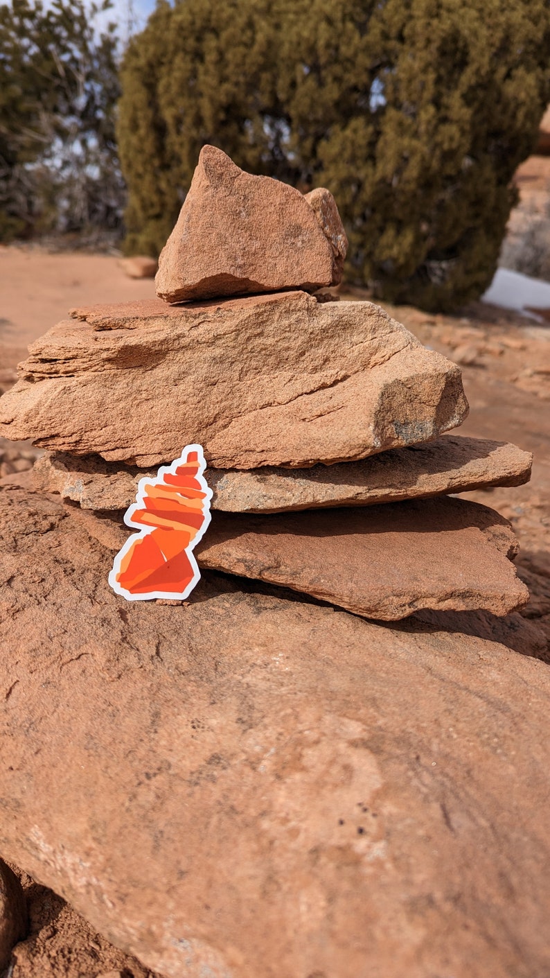 May include: A red and orange rock formation sticker placed on a stack of brown rocks in a desert landscape.