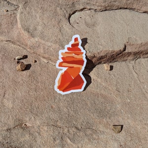 May include: A red and orange layered rock formation sticker. The sticker is on a brown, textured surface.