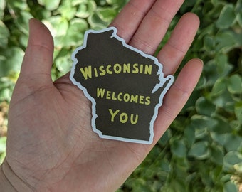 May include: A sticker in the shape of Wisconsin, with a dark brown background and a white border. The text "Wisconsin Welcomes You" is in green and yellow. The sticker is held in a hand against a background of green foliage.
