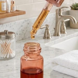 May include: A clear glass bottle with a rose gold dropper is held above a matching bottle filled with a reddish-brown liquid. Several drops of the liquid are visible on the counter. Other skincare products are in the background.