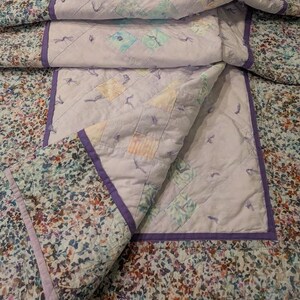 Puede incluir: Un edredón hecho a mano con un borde de estampado floral y un centro de color lila claro. El centro presenta patrones de diamantes y cuadrados en colores pastel. El edredón está doblado, mostrando las intrincadas costuras y el diseño.