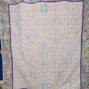 Może przedstawiać: Ręcznie robiona kołdra z jasnofioletowym środkiem z powtarzającym się wzorem kolorowych kwadratów i kwiatowych wzorów. Kołdra ma ciemnofioletową obwódkę i wzorzystą krawędź z mieszanką kolorów.