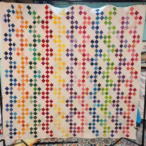 Puede incluir: Un edredón hecho a mano con un fondo color crema y un patrón repetido de formas de diamantes coloridos. Los diamantes están dispuestos en filas diagonales, creando un efecto vibrante de arcoíris. El edredón se exhibe en un soporte.