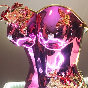 May include: A decorative, metallic pink bust sculpture with gold trim. The sculpture is adorned with jeweled butterflies and floral accents in shades of red, pink, and clear crystals. The base is black.