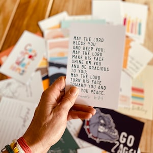 May include: A white card with a green inspirational quote about blessings and peace. The quote is from Numbers 6:24. The card is being held by a hand with a colorful bracelet.