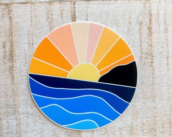 May include: A round sticker featuring a stylized sunset over the ocean. The design includes a yellow sun, orange and pink rays, a black mountain, and blue waves. The sticker is on a light wood surface.