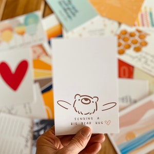 May include: A white greeting card with a brown bear illustration and the text "Sending a big bear hug" with a pink heart. The card is held in front of a collection of other cards with various designs and colors.