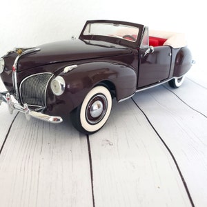 May include: A dark brown vintage convertible car model with white-walled tyres and chrome accents. The interior features red seating and a white convertible top. The car is a classic design, showcasing a detailed replica of a vintage automobile.