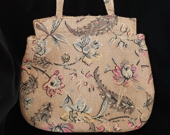 May include: A vintage handbag with a floral tapestry design. The bag features a beige background with colorful flowers and leaves in shades of pink, blue, and green. It has two handles and a structured shape, perfect for carrying essentials.
