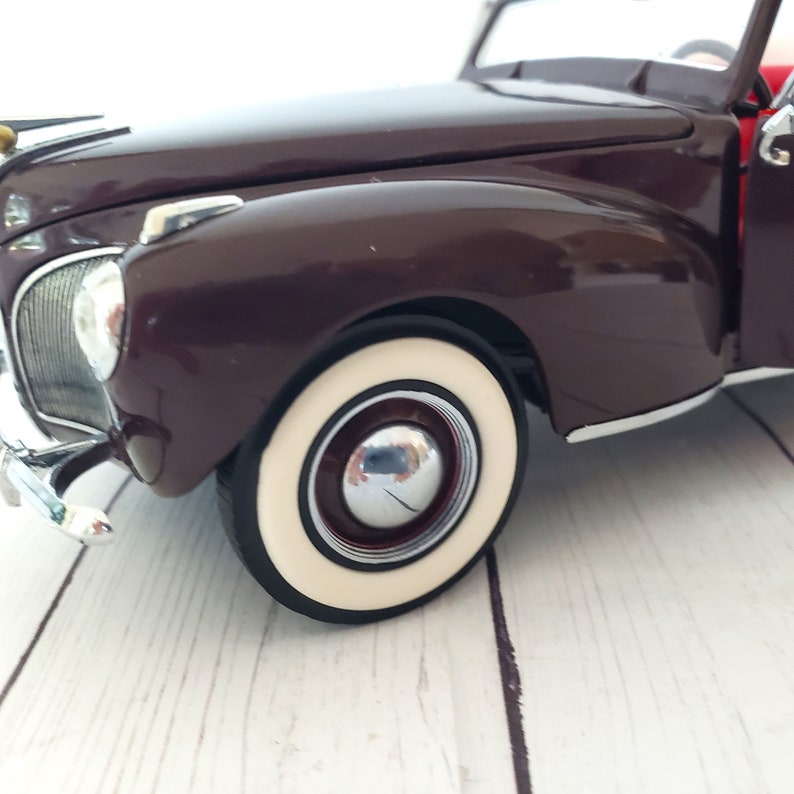 May include: A maroon classic car with a white-walled tyre. The car has chrome accents and a red interior. The tyre is approximately 25 cm in diameter. The car's door is open, revealing the interior.