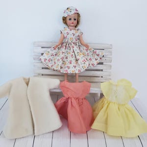 May include: A vintage doll wearing a floral print dress and matching hat, standing on a white bench. Additional doll clothing includes a cream-colored coat, a peach-colored dress, and a yellow dress with lace detailing. The items are arranged on a white wooden surface.