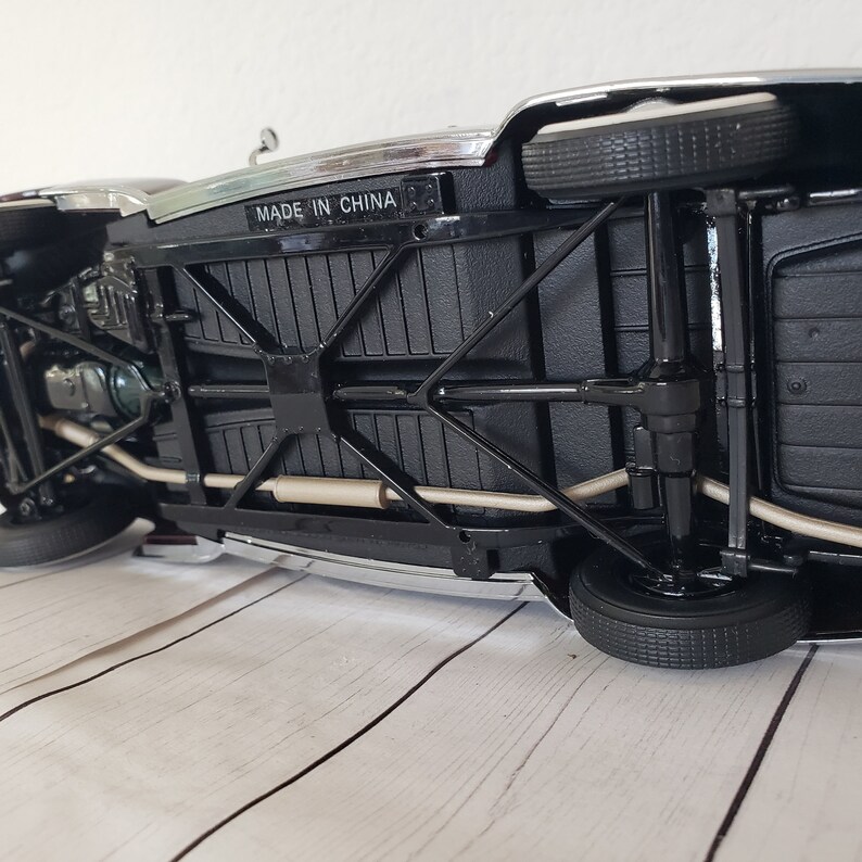 May include: Black die-cast model car with chrome detailing, visible undercarriage, and black tyres. The text "MADE IN CHINA" is visible on the underside. The car is on a light wood-grain surface.