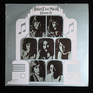 May include: Album cover for Three Dog Night's "Harmony" album. The cover features a silver and white design with the band's name and album title at the top. Portraits of the band members are arranged in a grid, with musical notes as accents.