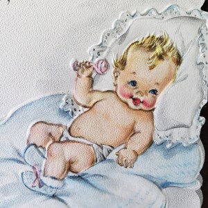 Op de afbeelding: Vintage baby-thema wenskaart met een cherubijnachtige baby met blond haar, blauwe ogen en roze wangen. De baby ligt op een blauwe deken en kussen en houdt een roze rammelaar vast. De kaart heeft een getextureerd oppervlak.