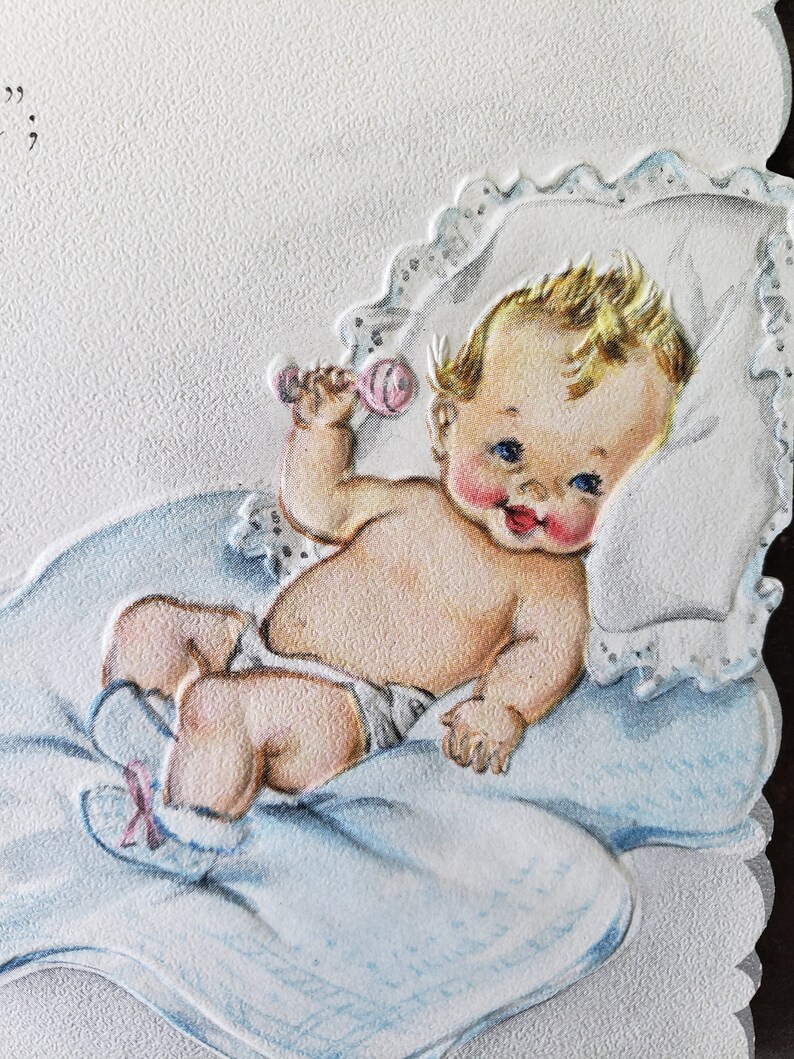 Op de afbeelding: Vintage baby-thema wenskaart met een cherubijnachtige baby met blond haar, blauwe ogen en roze wangen. De baby ligt op een blauwe deken en kussen en houdt een roze rammelaar vast. De kaart heeft een getextureerd oppervlak.