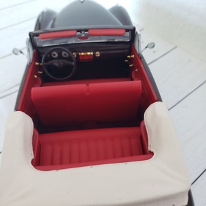 May include: A black convertible car model with a red interior. The model features a black steering wheel, dashboard, and side mirrors. The convertible top is off-white.