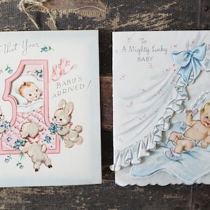Op de afbeelding: Twee vintage baby wenskaarten. E&eacute;n kaart zegt "Now That Your Baby's Arrived!" met een baby in een roze frame, omringd door teddyberen en konijntjes. De andere kaart zegt "To A Mighty Lucky Baby" met een baby in een wieg.