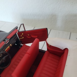 May include: A detailed interior of a black convertible car model. The car has red seats, a black dashboard, and gold-coloured accents. The door is open, revealing the interior details. The convertible top is white.