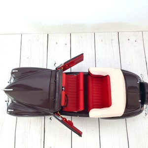 May include: A detailed model of a classic convertible car. The car is a deep brown colour with red interior seating and a cream-coloured convertible top. The doors are open, showcasing the interior details.