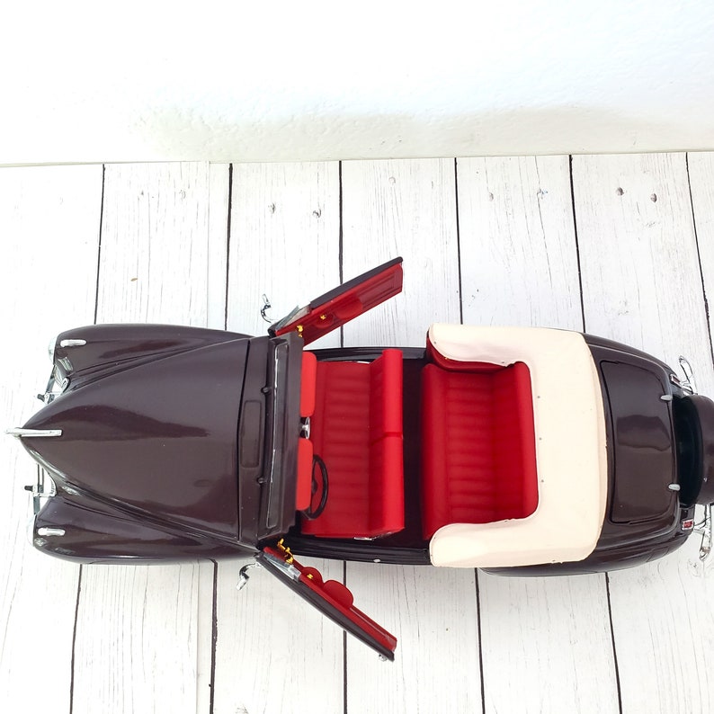 May include: A detailed model of a classic convertible car. The car is a deep brown colour with red interior seating and a cream-coloured convertible top. The doors are open, showcasing the interior details.