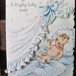 Op de afbeelding: Vintage baby wenskaart met een baby illustratie. De kaart toont een baby in een blauwe deken, die een rammelaar vasthoudt. De tekst luidt "To A Mighty Lucky BABY" met een blauwe strik en bloemaccenten.