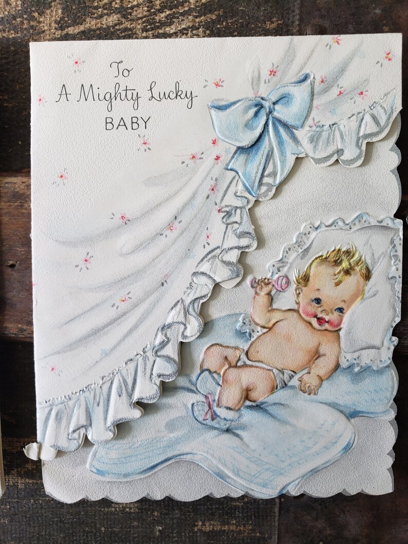 Op de afbeelding: Vintage baby wenskaart met een baby illustratie. De kaart toont een baby in een blauwe deken, die een rammelaar vasthoudt. De tekst luidt "To A Mighty Lucky BABY" met een blauwe strik en bloemaccenten.