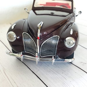 May include: A maroon vintage car model with chrome detailing on the bumper, headlights, and grille. The car features a white convertible top and a red interior. This is a classic car model.
