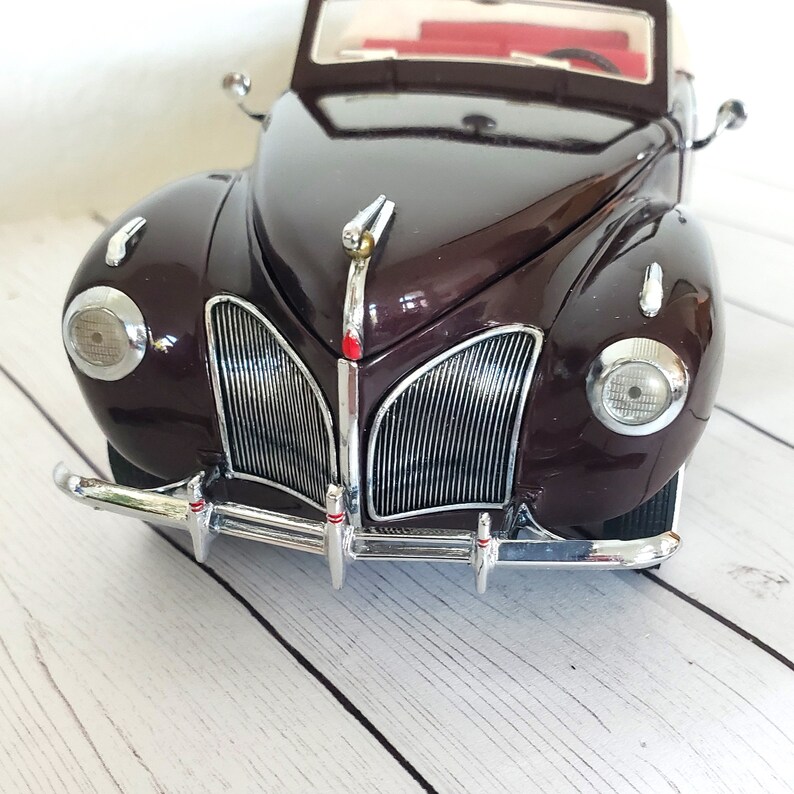 May include: A maroon vintage car model with chrome detailing on the bumper, headlights, and grille. The car features a white convertible top and a red interior. This is a classic car model.