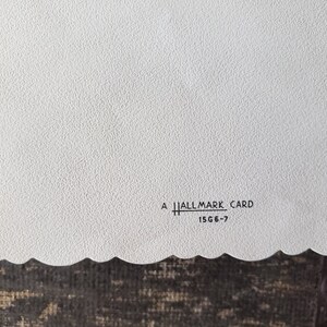 Op de afbeelding: Een witte Hallmark-kaart met een getextureerd oppervlak en een geschulpte rand. De tekst "A Hallmark Card" en "15G6-7" is zwart gedrukt. De kaart ligt op een donkerbruin oppervlak.