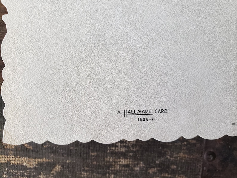 Op de afbeelding: Een witte Hallmark-kaart met een getextureerd oppervlak en een geschulpte rand. De tekst "A Hallmark Card" en "15G6-7" is zwart gedrukt. De kaart ligt op een donkerbruin oppervlak.