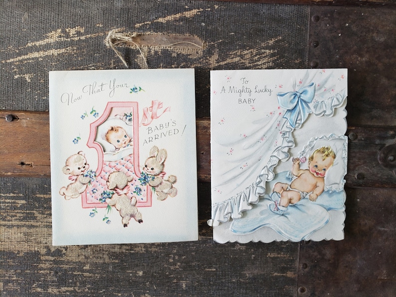 Op de afbeelding: Twee vintage baby-wenskaarten. E&eacute;n kaart zegt "Now That Your Baby's Arrived!" met een baby in een wieg en dierlijke illustraties. De andere kaart zegt "To A Mighty Lucky Baby" met een baby in een deken.