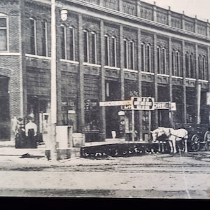 May include: Black and white vintage photograph of a street scene with buildings and horse-drawn carriages. A sign reads "PIANOS" on the corner building. Other signs are visible, including one for a "DRUG STORE".