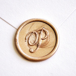 Puede incluir: Un sello de cera dorado con las letras "OP" en una elegante escritura, adherido a un sobre blanco. El sello es redondo y tiene una apariencia texturizada, lo que sugiere una estética vintage o artesanal. El fondo es de color blanco roto.