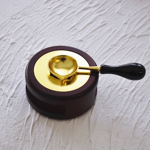 May include: A gold-colored wax seal spoon with a black wooden handle sits on a dark brown wooden base with a gold-colored top.