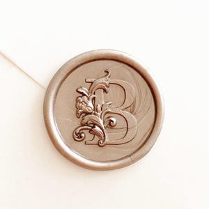 May include: A metallic silver wax seal with the letter "B" in a serif font, adorned with floral embellishments. The seal is circular and sits on a white surface, suggesting stationery or crafting supplies.