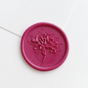 May include: A round, magenta wax seal with a floral design. The seal is embossed with a detailed flower, featuring delicate petals and stems. The seal is set against a white background, creating a contrast that highlights the seal's intricate details.