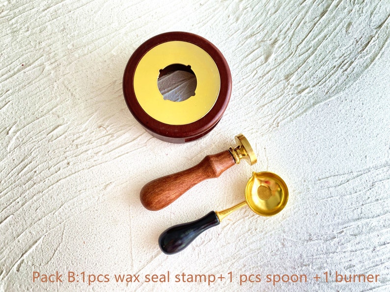 May include: A wax seal stamp set with a wooden handle, a gold-colored spoon, and a burner. The set includes a round, dark brown wax seal stamp with a gold-colored interior. The spoon has a gold bowl and a black handle. The text "Pack B" is visible.