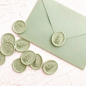 May include: Sage green wax seals with various embossed designs, including a Christmas tree and floral sprigs, are scattered around a matching envelope. The seals are oval and circular, adding a decorative touch to stationery or crafts.