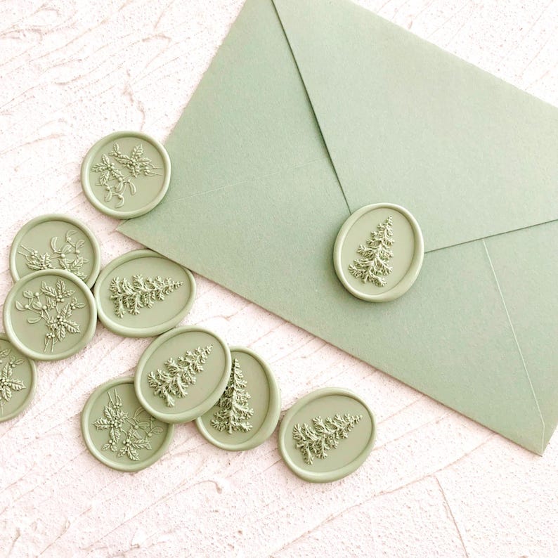 May include: Sage green wax seals with various embossed designs, including a Christmas tree and floral sprigs, are scattered around a matching envelope. The seals are oval and circular, adding a decorative touch to stationery or crafts.