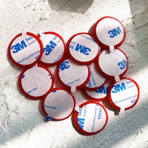 May include: A collection of red and white 3M double-sided adhesive tape circles. Each circle has a white center with blue 3M branding and product information. The red edges are visible, and the tape is designed for double-coated tissue applications.