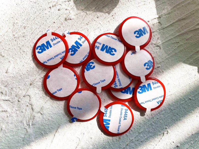 May include: A collection of red and white 3M double-sided adhesive tape circles. Each circle has a white center with blue 3M branding and product information. The red edges are visible, and the tape is designed for double-coated tissue applications.