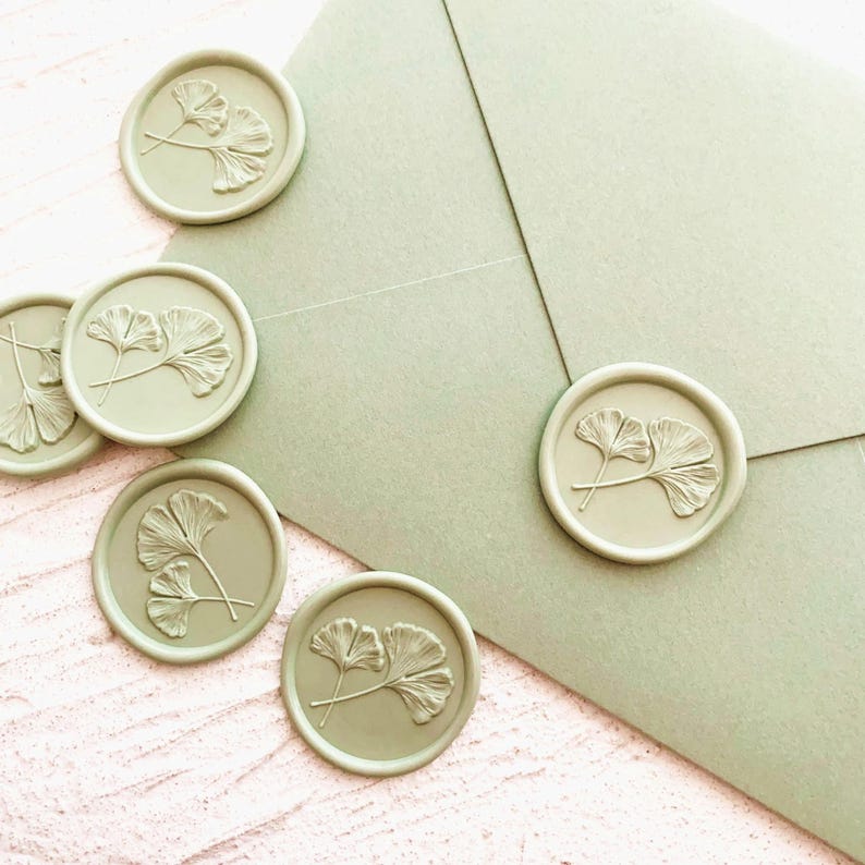 May include: Sage green wax seals with a ginkgo leaf design are scattered around a matching envelope. The seals are round and embossed with the delicate leaf pattern. The envelope is a soft, muted green.