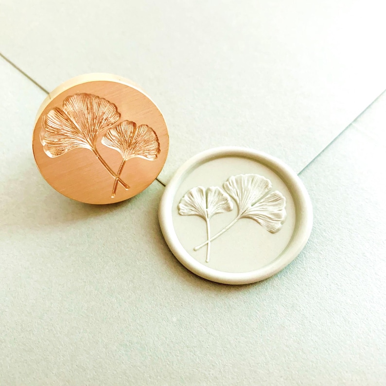 May include: A copper-colored wax seal stamp and a white wax seal with a detailed ginkgo leaf design. The stamp is round, and the leaves are crossed. The white seal is on a light blue surface.