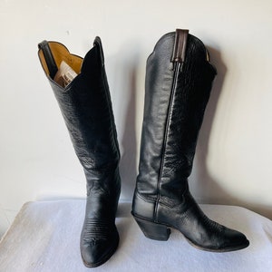 Puede incluir: Un par de botas de vaquero de cuero negro. Las botas tienen un acabado texturizado, puntera en punta y costuras decorativas en el pie. Las cañas son altas, con un borde superior festoneado. Los tacones miden unos 5 cm de alto.