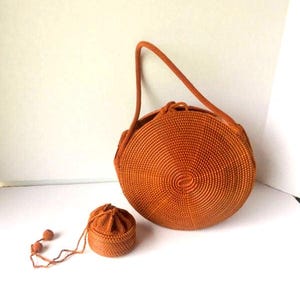 May include: A round, orange handbag with a matching small drawstring pouch. The handbag has a handle and is made of woven material. The pouch has a drawstring closure and two decorative beads. The items are set against a white background.