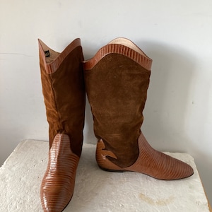 Könnte beinhalten: Ein Paar braune Lederstiefel mit einem Obermaterial aus Wildleder. Die Stiefel haben ein strukturiertes, reptilienartiges Design am unteren Teil und einen dekorativen Ausschnitt an der Seite. Die Stiefel sind kniehoch und haben eine flache Sohle.